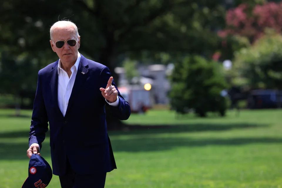 A tieless President Biden, wearing trademark aviator sunglasses and holding a blue cap in his right hand, walks on the South Lawn of the White House.